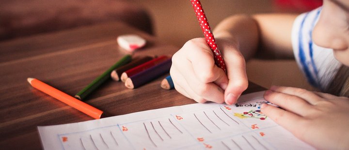 Kind mit Bleifstift in der Hand und Buntstiften auf dem Tisch über Arbeitsblatt